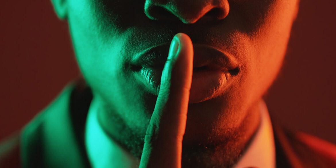 A person in sunglasses holds a finger to their lips under green and orange lighting.