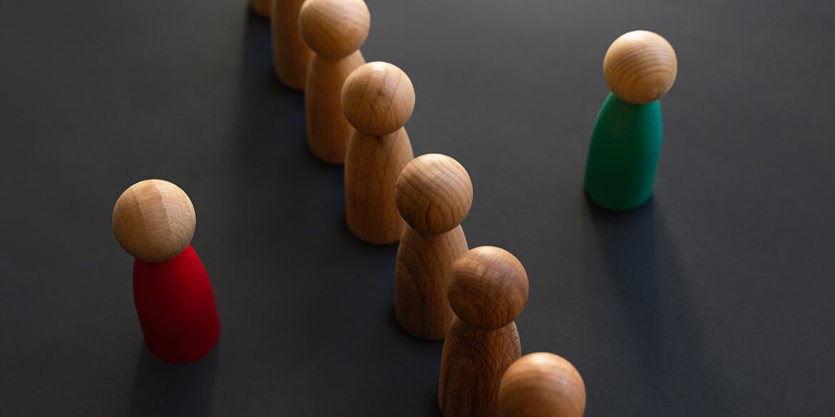 A red wooden figure and a green wooden figure stand on opposite sides of a row of unpainted wooden figures.